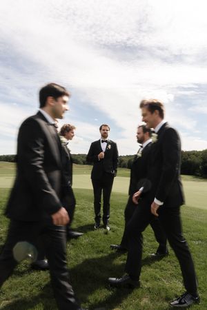 Groom standing with groomsmen in black tuxedos on a sunny green outdoor field before the wedding.