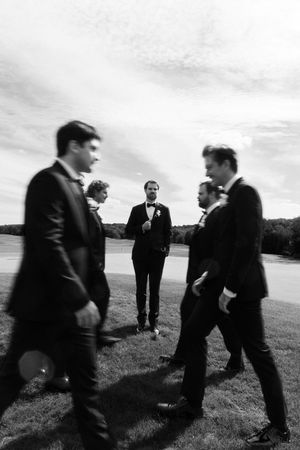 Black and white photo of groomsmen in tuxedos walking outdoors on grass with a pond in the background.