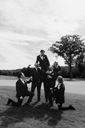 Groomsmen in black tuxedos pose playfully outdoors, lifting the groom in a celebratory wedding photo.