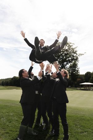 Groomsmen in black suits toss a laughing groom into the air on a green golf course at a wedding celebration.
