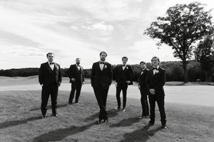Group of groomsmen in black tuxedos posing outdoors on a golf course.