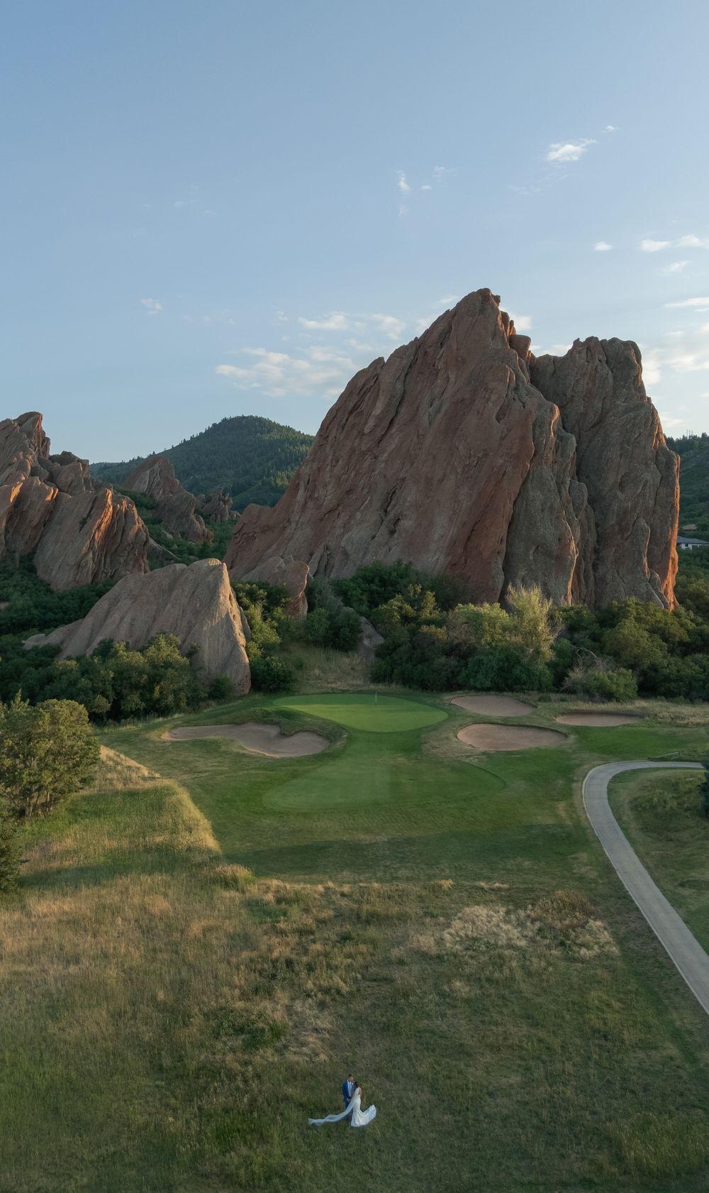 Aerial view of a golf course green and sand bunkers beneath towering red sandstone rock formations, with a couple in wedding attire below.