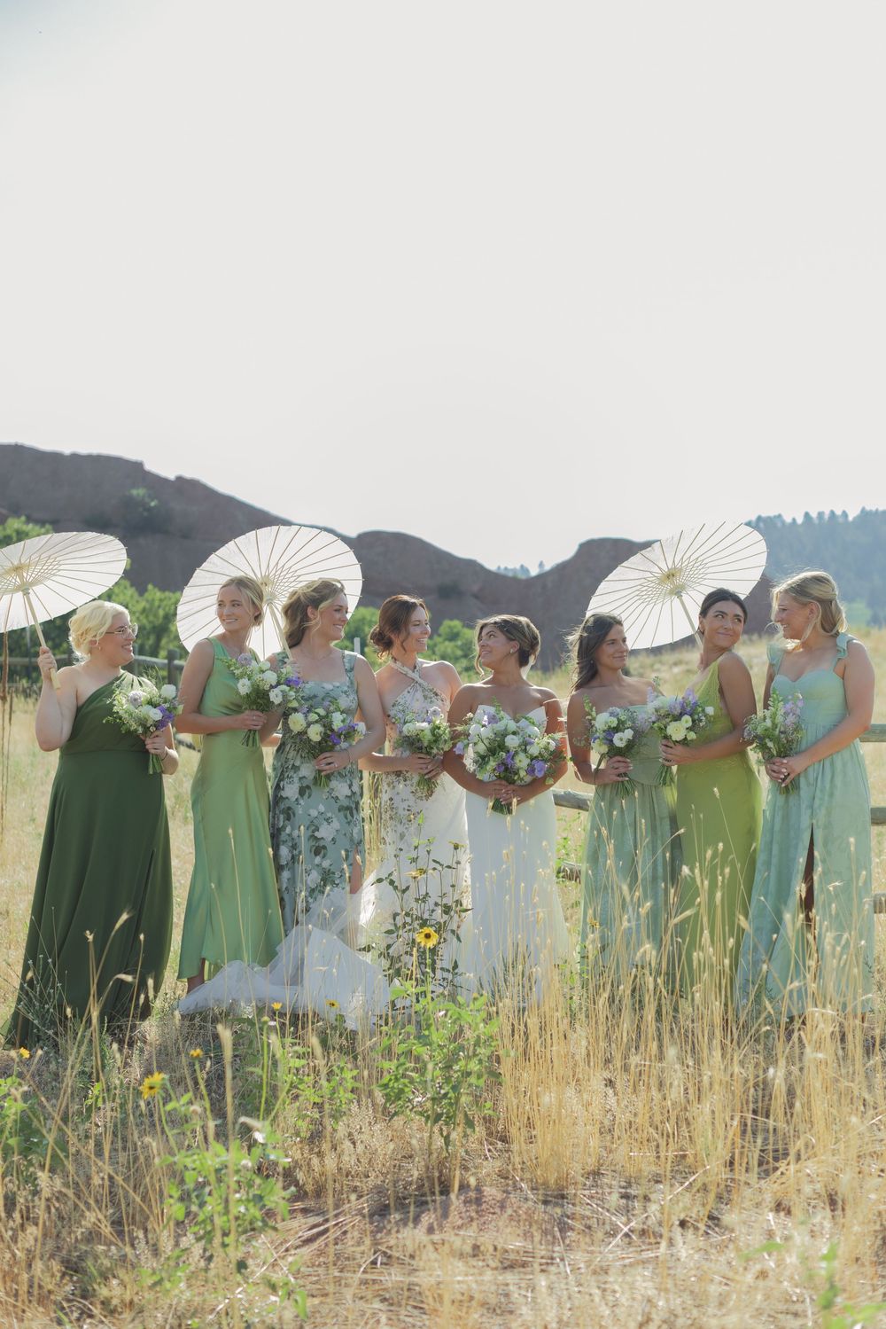 Bride in white gown stands among bridesmaids in mixed green dresses holding bouquets and white parasols in a sunny mountain field.