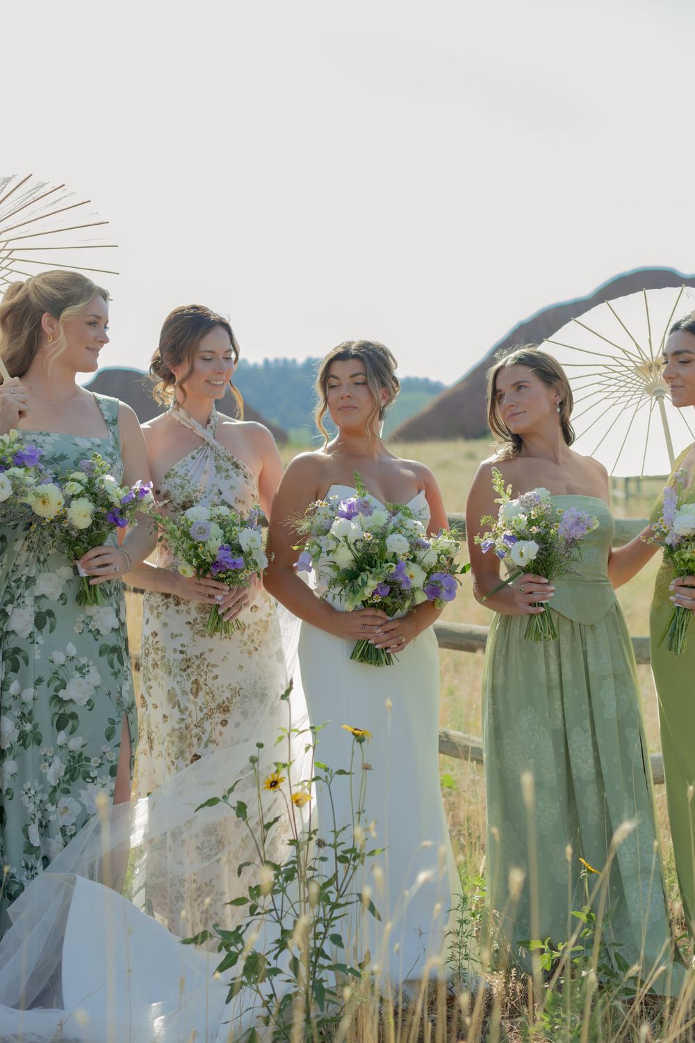Bride and bridesmaids in sage green and floral dresses hold pastel bouquets in a sunny field with parasols and rolling hills behind them.