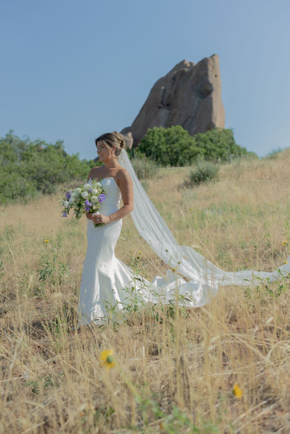 Bride in a white gown with a long flowing veil holds a bouquet while walking through golden grass with a red rock formation behind her.