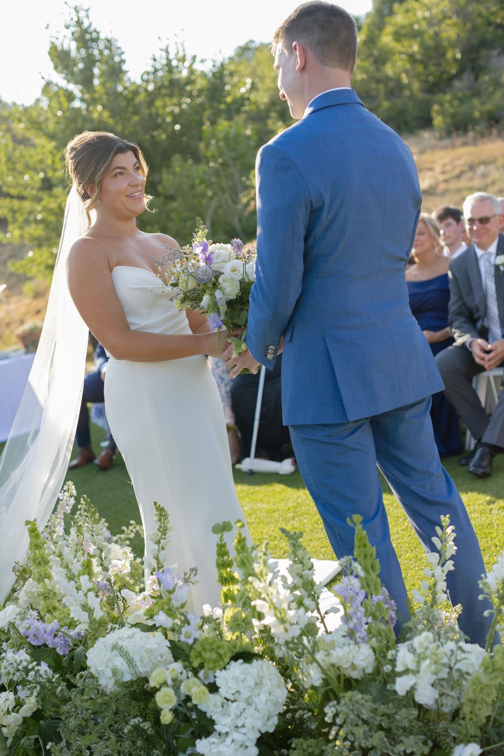 Bride in a strapless white gown and veil holds a bouquet while facing a groom in a blue suit at an outdoor garden wedding ceremony.