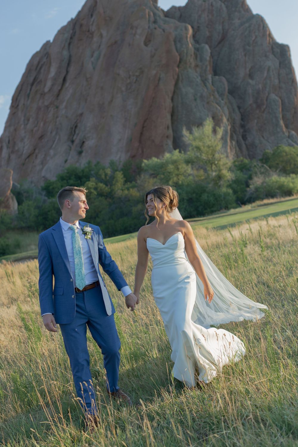Bride in a white gown with a flowing veil holds hands with a groom in a blue suit while walking through a grassy field near red rock formati