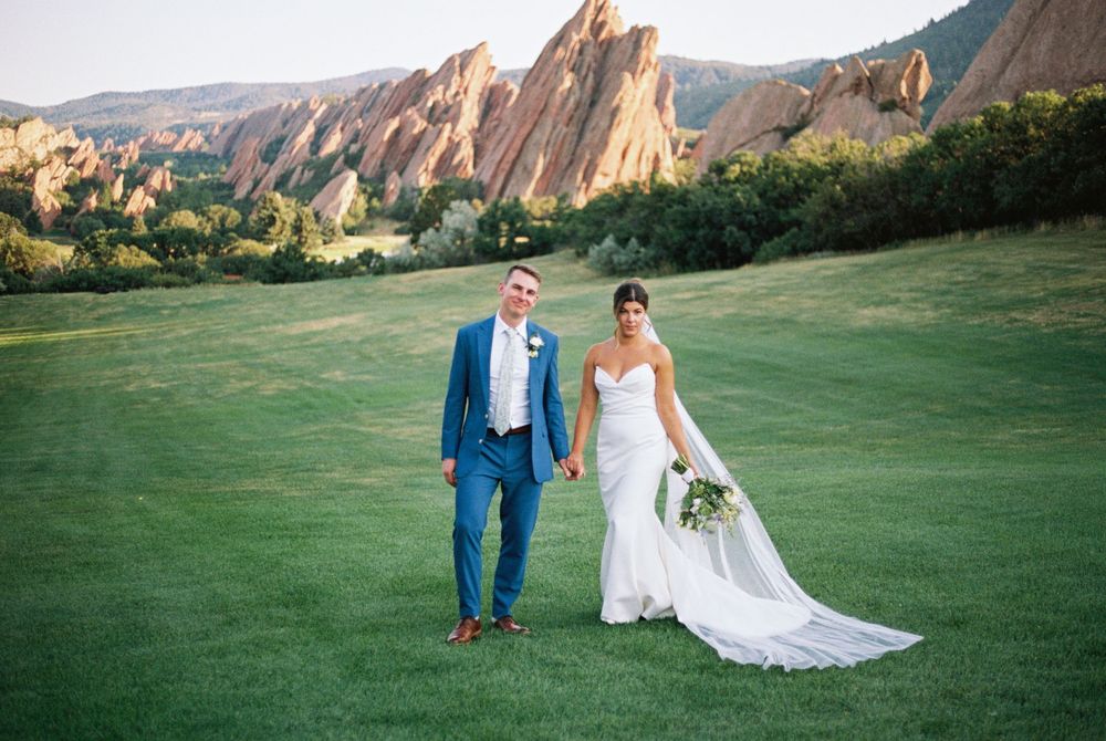 Bride in a white gown and groom in a blue suit hold hands on a green lawn with red sandstone rock formations behind them.