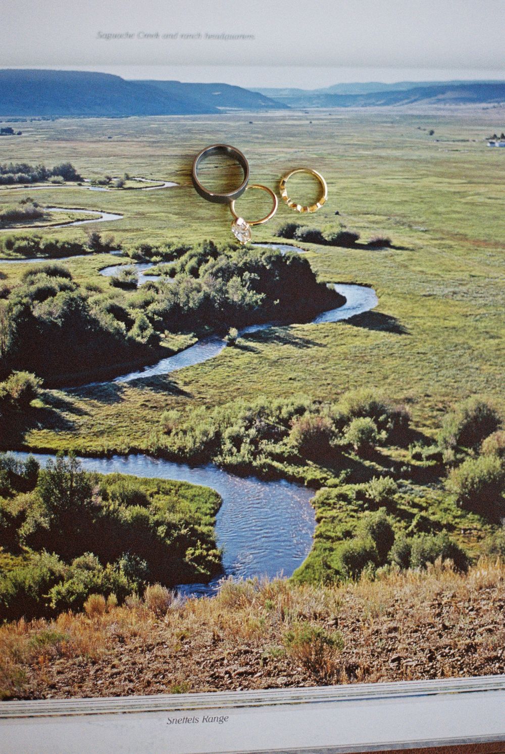 Wedding rings resting on a printed photo of a winding river curving through a lush green valley meadow with distant mountains.