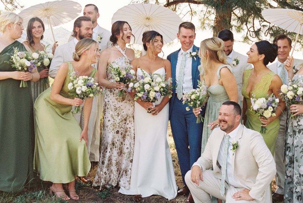 Smiling bride in a white gown holding a bouquet, surrounded by bridesmaids in sage green dresses and groomsmen under umbrellas outdoors.