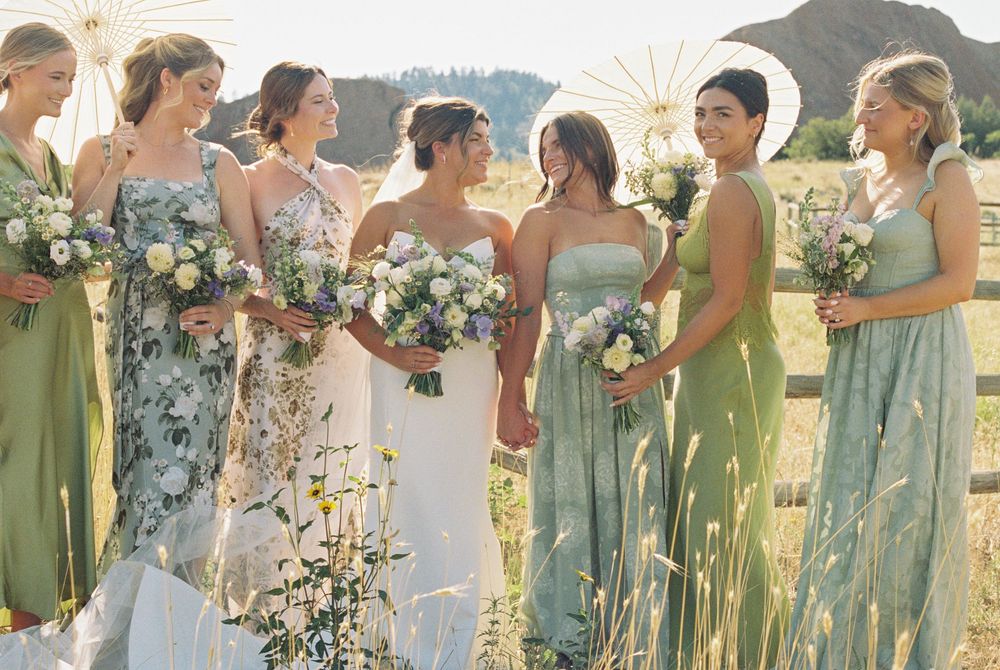 Bride in white gown surrounded by bridesmaids in sage green dresses holding wildflower bouquets in a sunny field with mountains behind.