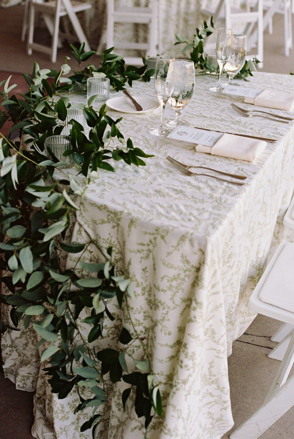 Wedding reception table with a green and white botanical linen, eucalyptus garland, folded napkins, place settings and crystal glassware.