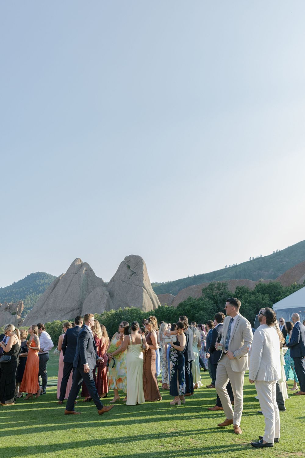 Wedding guests in formal attire mingle on a sunny green lawn with red rock formations and mountains in the background.