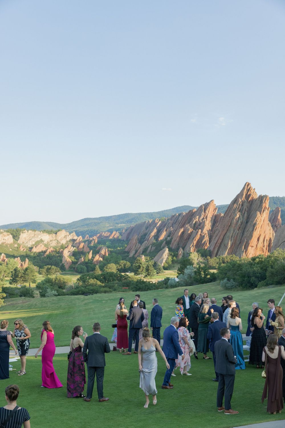 Wedding guests in formal attire mingle on a green lawn with red sandstone rock formations and mountains in the background.