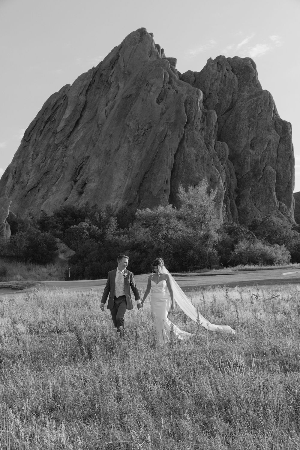 Bride and groom holding hands and walking through a grassy field in front of a massive rock formation in a black and white wedding photo.