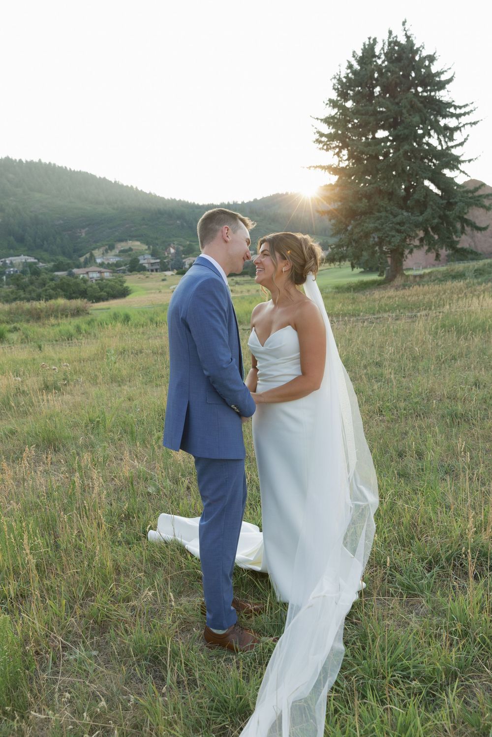 Bride in a white gown with a long veil and groom in a blue suit share an intimate moment in a grassy field at sunset with mountains behind.