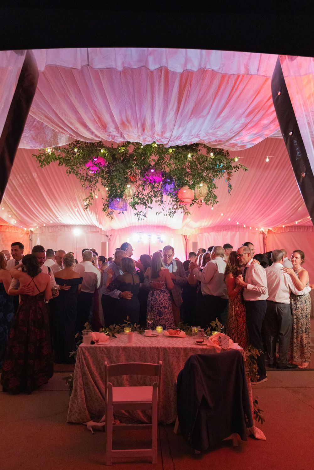 Wedding guests mingle under a draped pink tent with a hanging floral chandelier, string lights, and a candlelit reception table.
