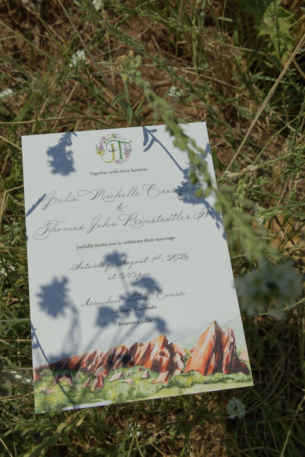 Watercolor wedding invitation with red rock formations and a monogram, resting in grass with wildflower shadows falling across the card.