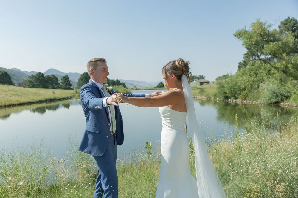 Bride in a white gown and veil holds hands with a groom in a blue suit beside a calm pond with grassy banks and mountains behind.
