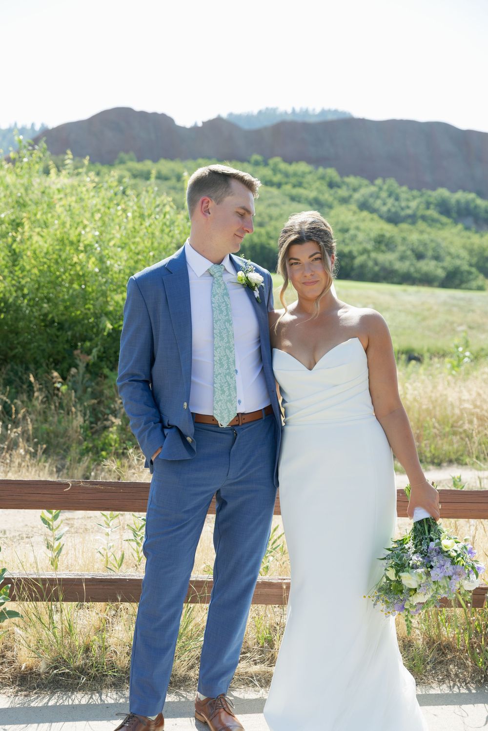 Smiling bride in a white gown holding a bouquet stands beside a groom in a blue suit near a wooden fence with red rock hills behind.