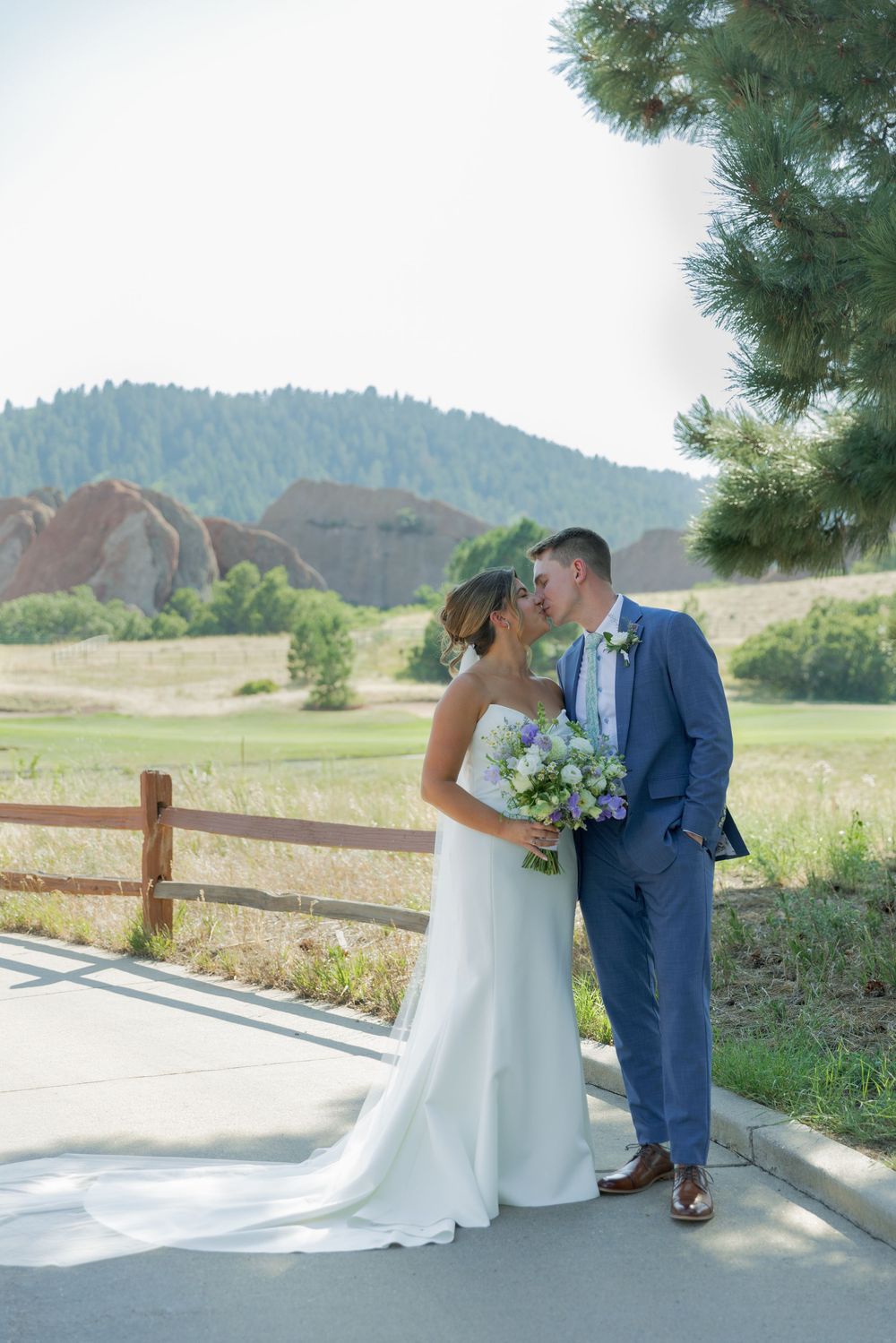 Bride in a white gown holding a bouquet kisses groom in a blue suit on a paved path with red rock formations and pine trees behind.