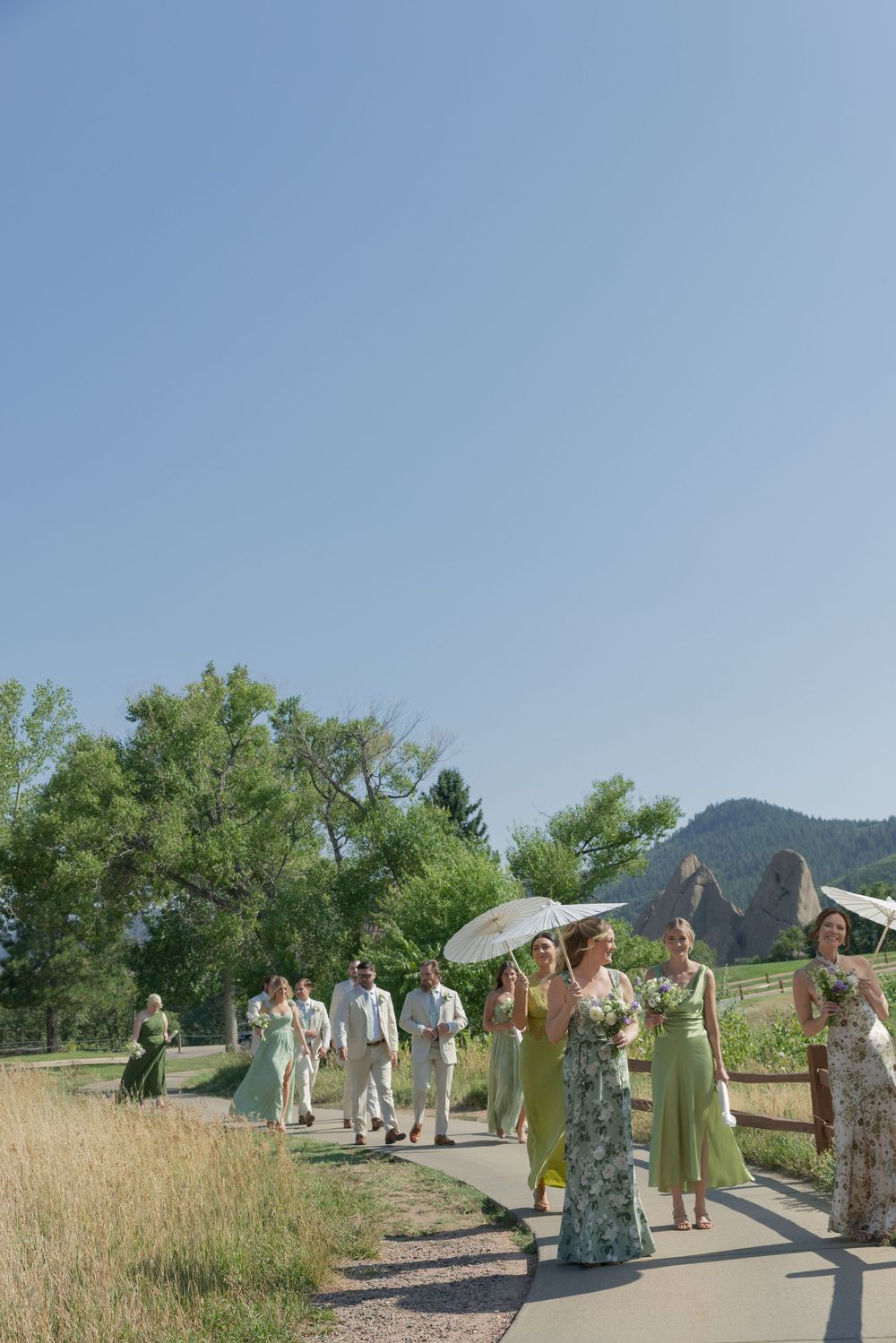 Wedding party in sage green dresses and light suits walks a paved path beneath white parasols with mountains and trees in the background.