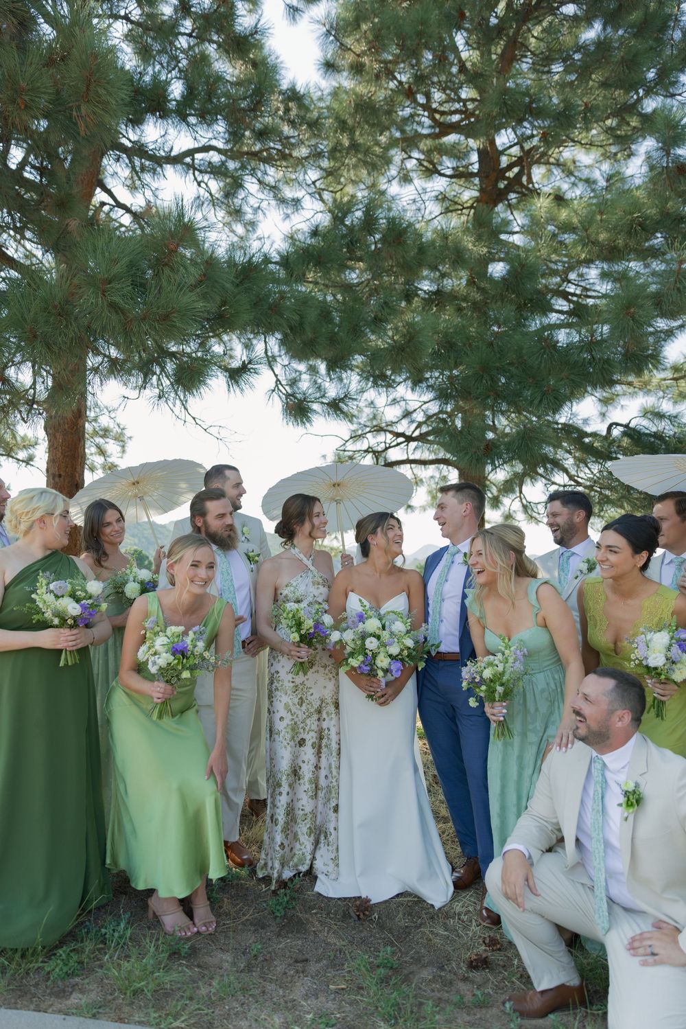 Bride in a white gown laughs with bridesmaids in green dresses and groomsmen holding bouquets beneath pine trees and white parasols.
