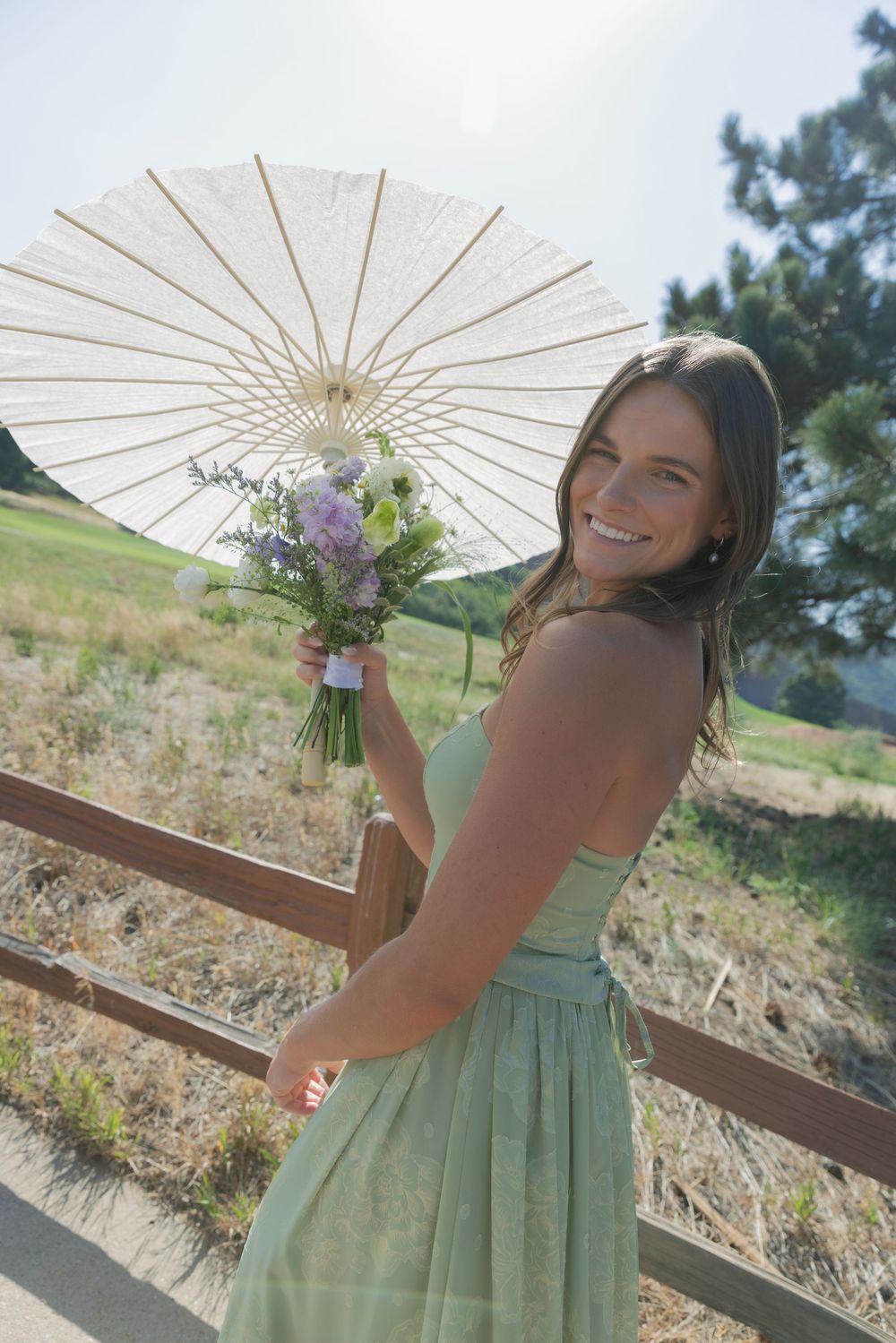 Smiling bridesmaid in a sage green dress holds a purple wildflower bouquet and white paper parasol beside a rustic wooden fence outdoors.