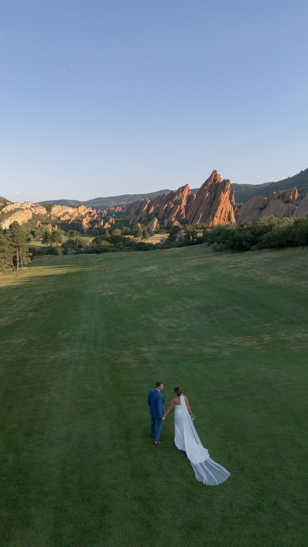 Bride in a flowing white gown and groom in a blue suit walk hand in hand across a green lawn toward red sandstone rock formations at sunset.