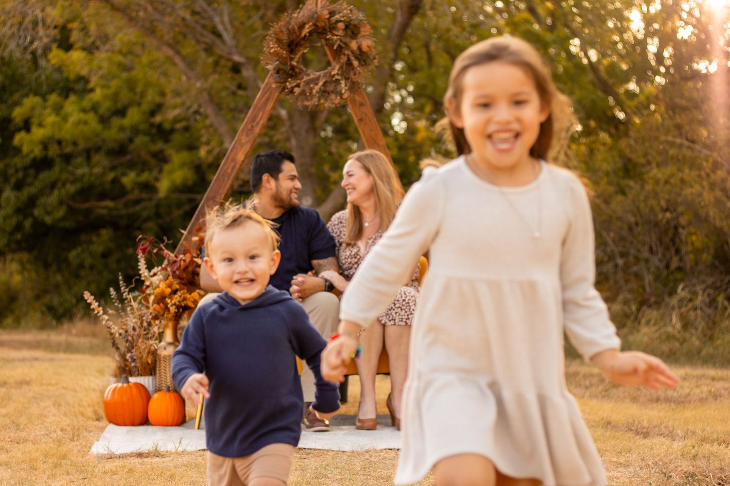 Family enjoying autumn outdoor photoshoot with pumpkins, two young children playing while parents sit on blanket.