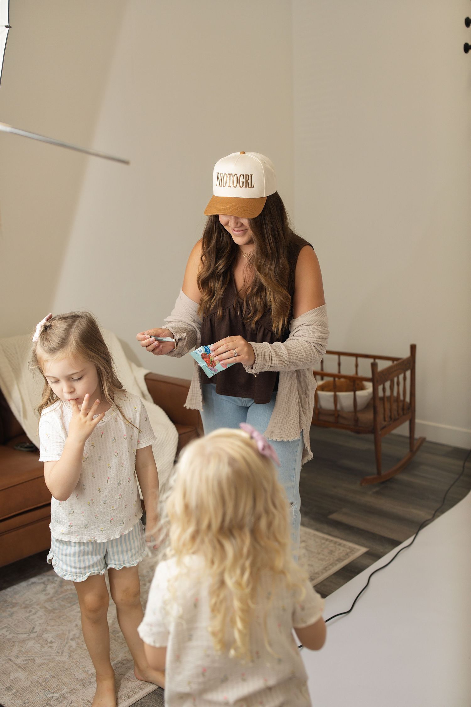 A mother sits on the floor using a laptop while three young girls play around her with toys and a baby doll.