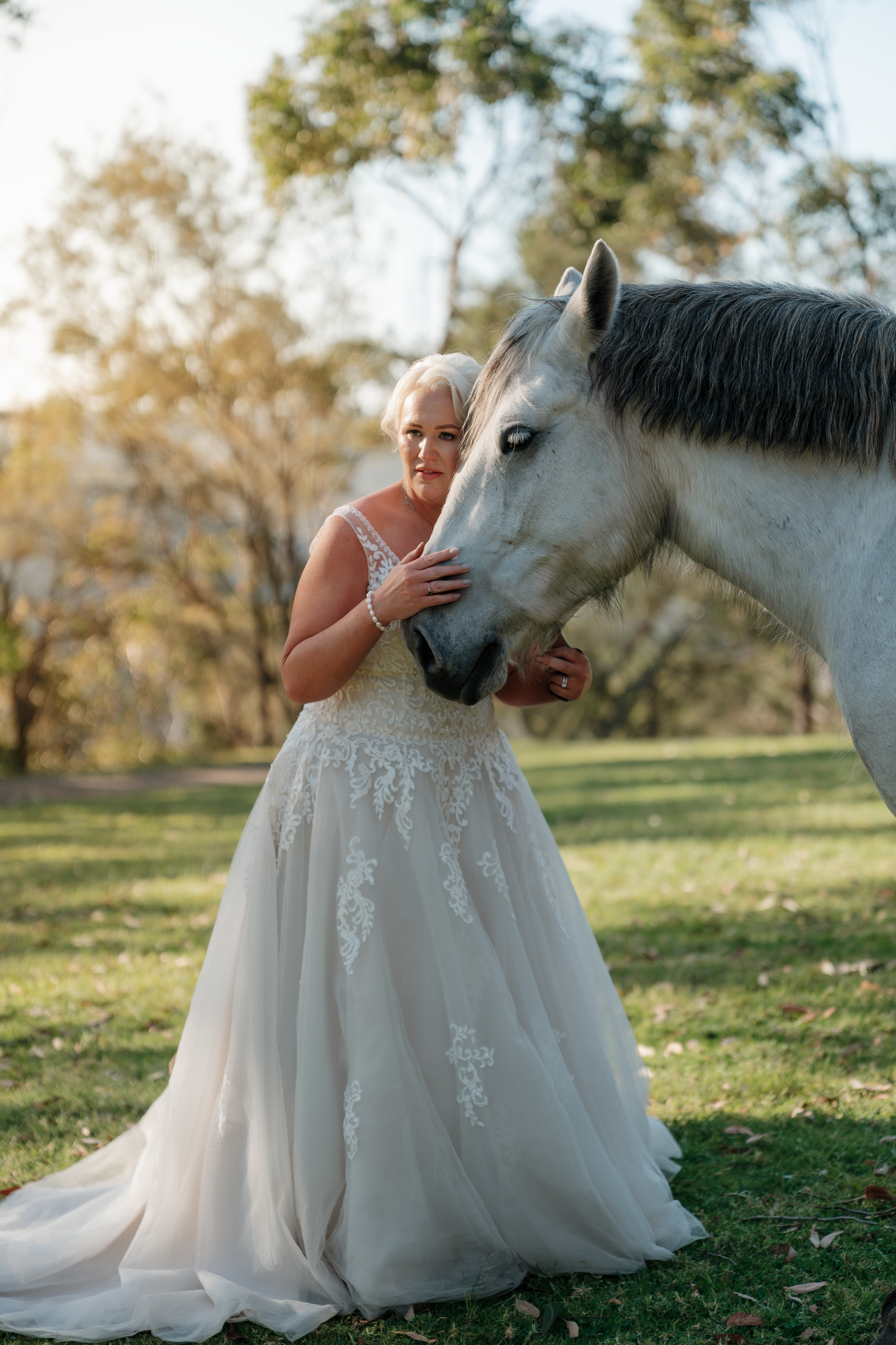 Bride in lace wedding gown tenderly holds the nose of a white horse in a sunlit green park.