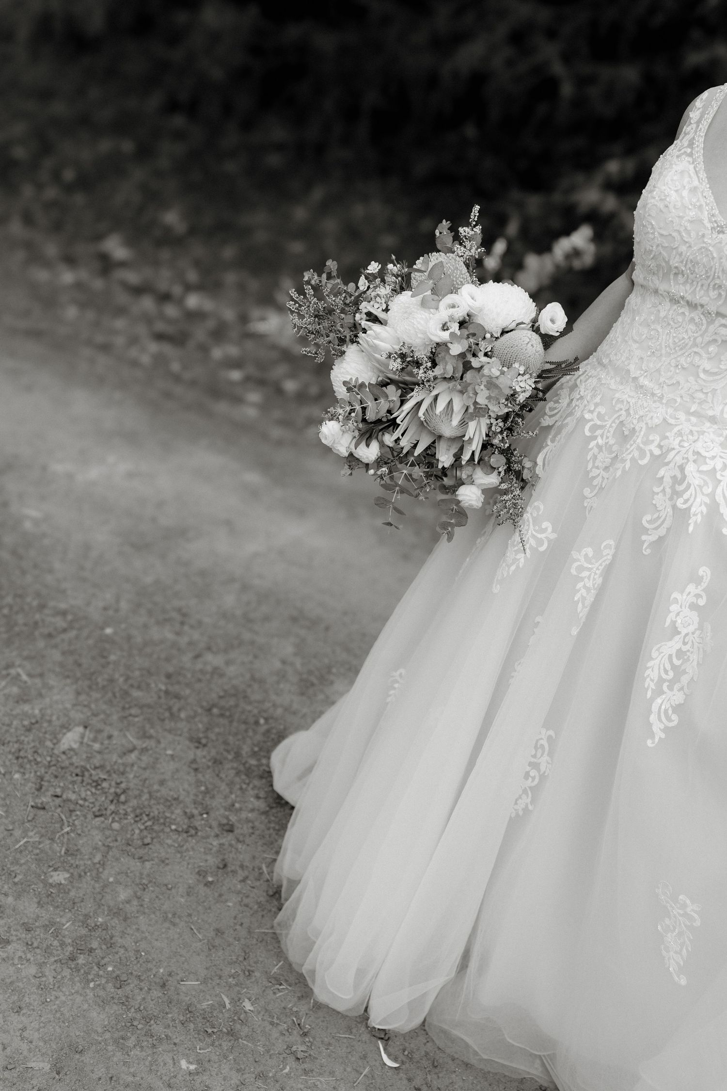 Black and white photo of a bride in lace gown holding a floral bouquet with peonies and greenery outdoors.