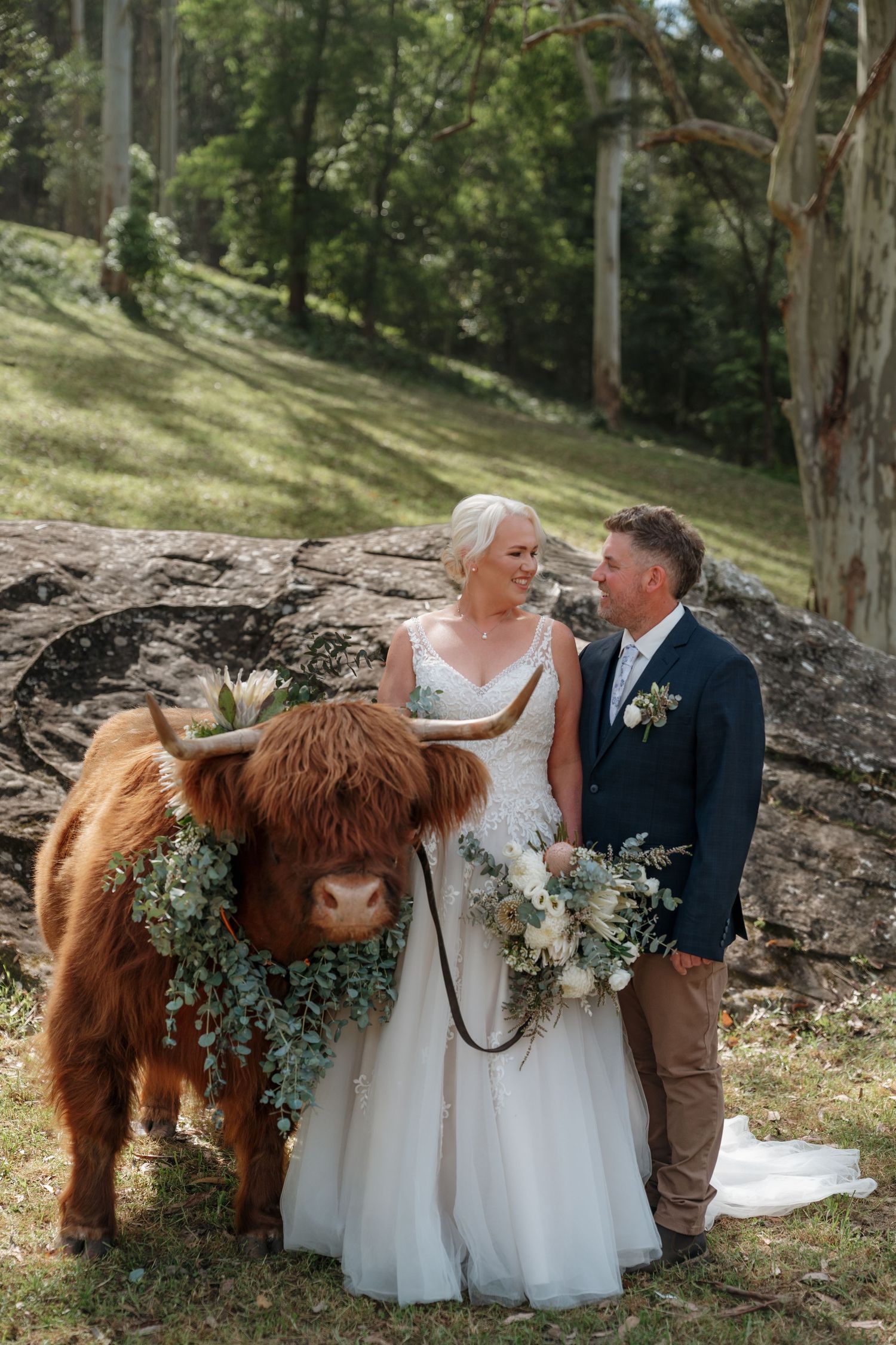 Bride and groom posing with a decorated Highland cow wearing floral garland at an outdoor rustic wedding ceremony.