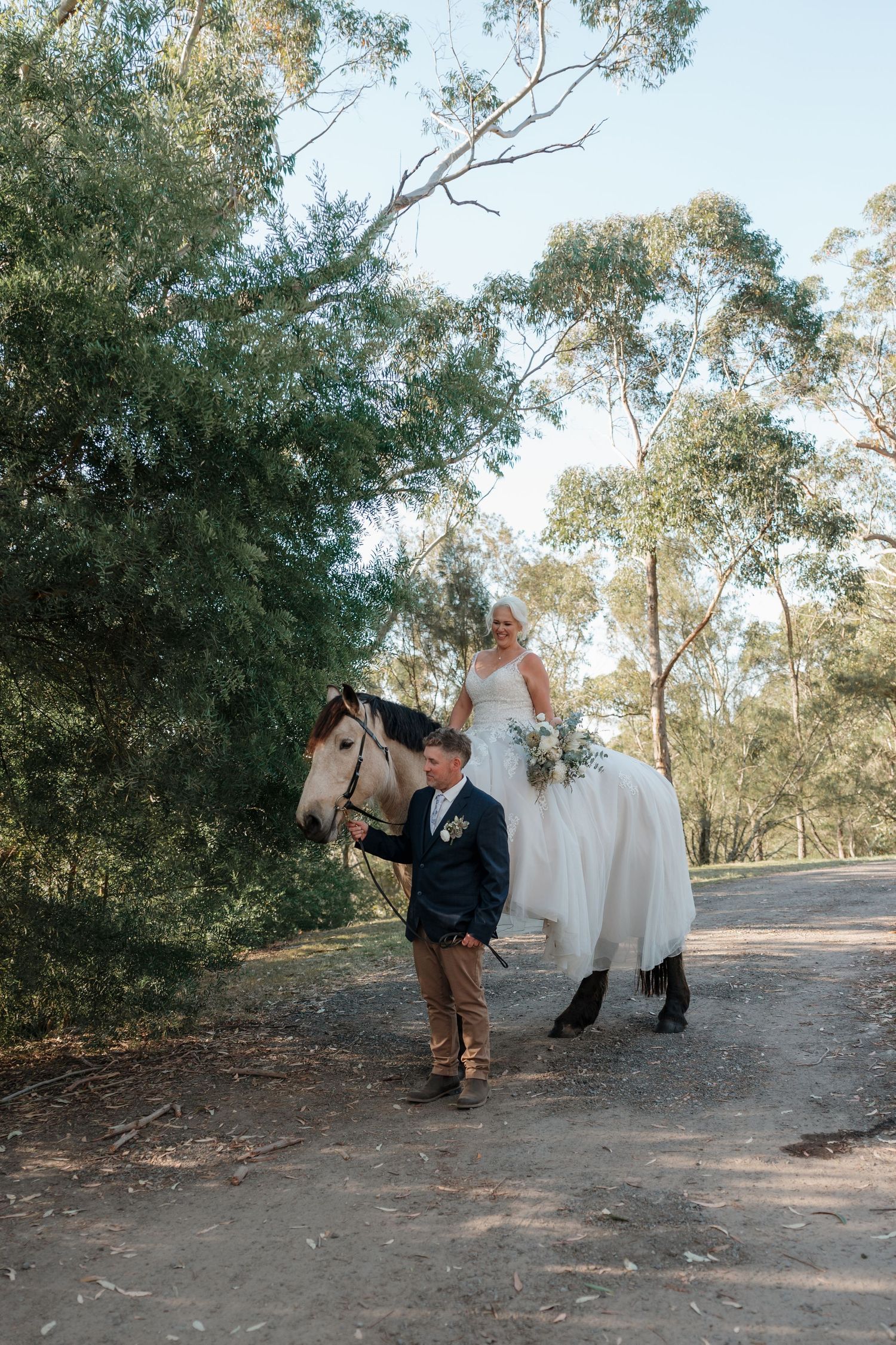 Bride in white gown rides white horse led by groom on a scenic bush path surrounded by eucalyptus trees.