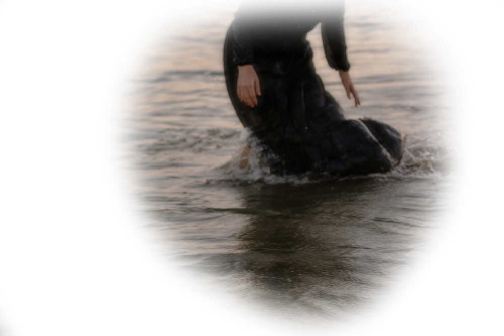 Woman in black wetsuit wading through shallow ocean water with vignette effect.