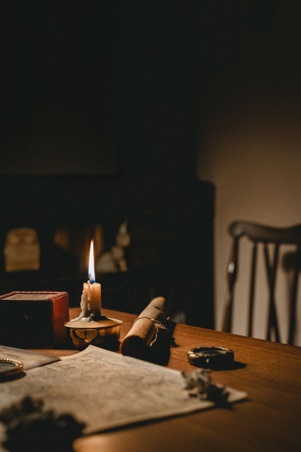 A dimly lit wooden table with a glowing candle, rolled scroll, old map, and antique objects in a moody, vintage setting.