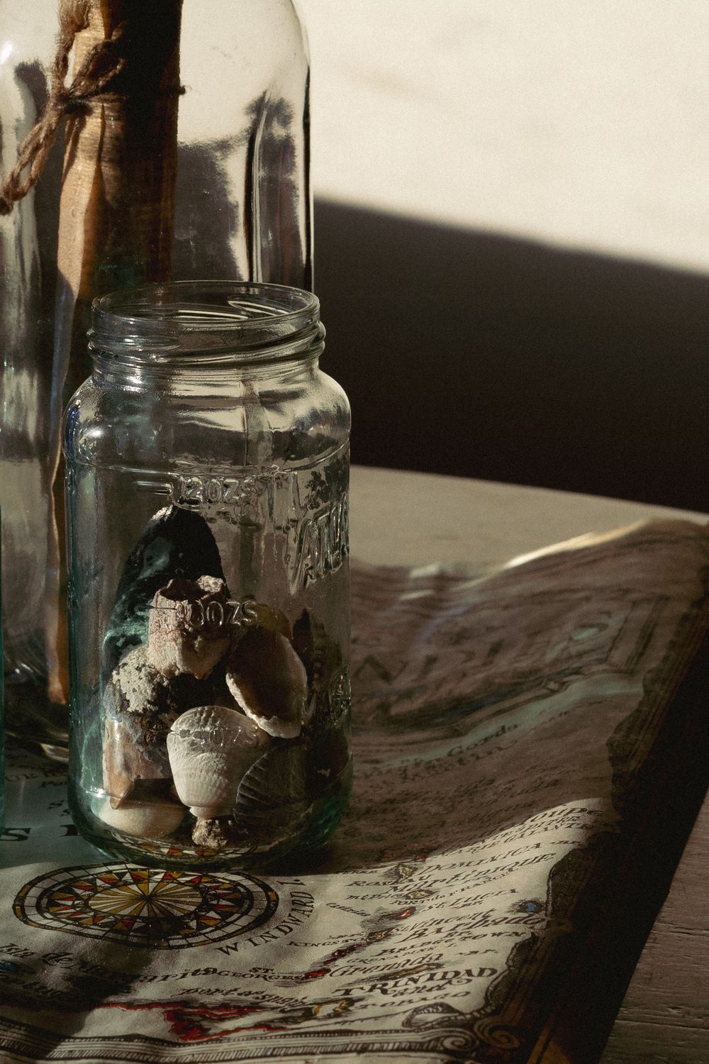 Glass mason jar filled with seashells beside a tall bottle on a vintage map, bathed in warm natural sunlight.