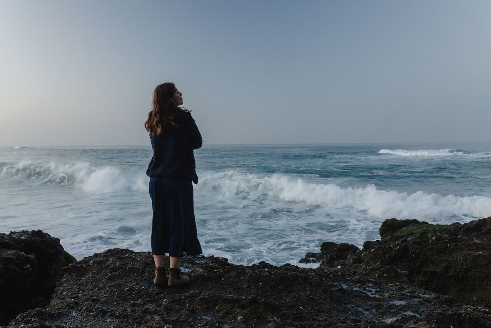 Woman standing on rocky shore of Victoria Beach Laguna Beach, California gazing at crashing ocean waves during dusk in a dark blue outfit.