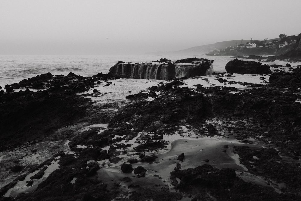 Black and white coastal landscape with rocky shoreline, tide pools, and misty ocean horizon.
