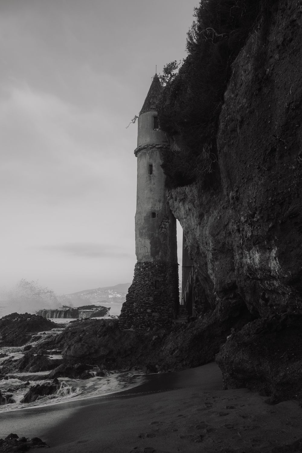 Black and white photo of an ancient stone tower built into coastal cliffs beside a sandy beach under a cloudy sky.