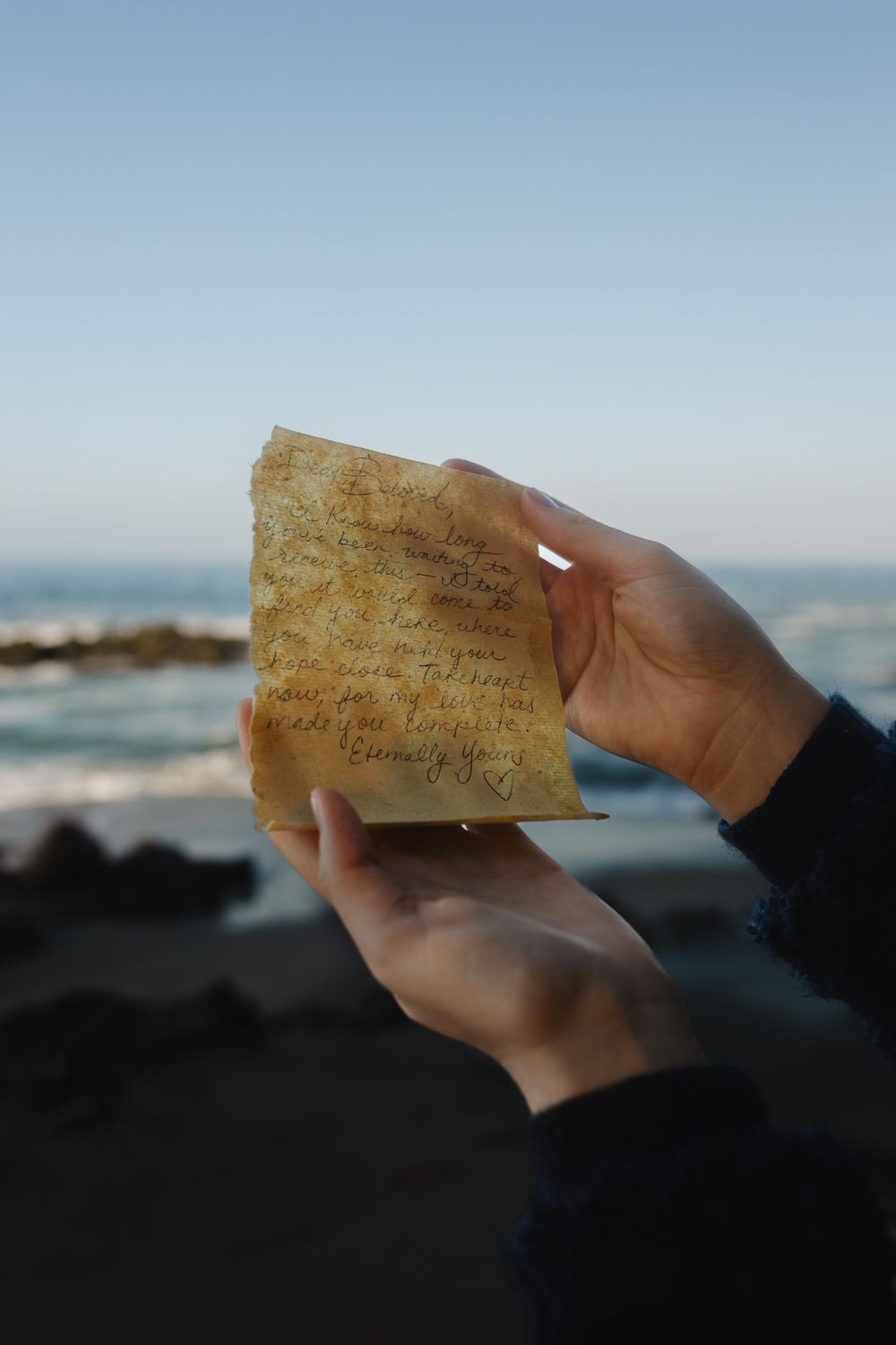Hands holding an aged handwritten letter on Laguna beach with California ocean waves and rocky shore in the background.