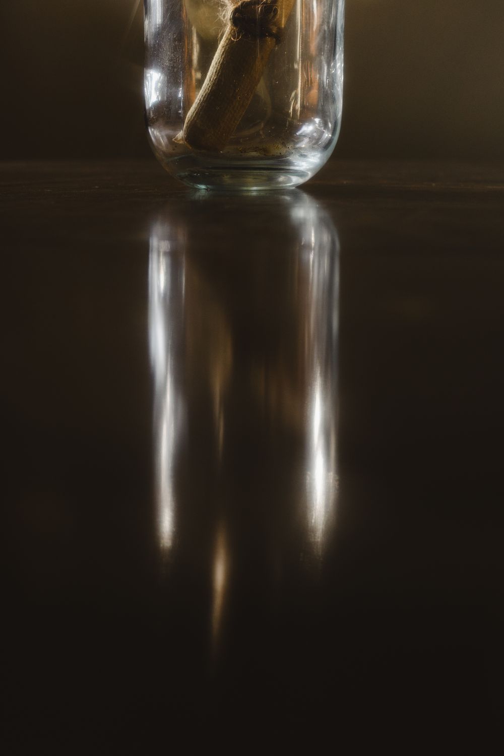 Glass jar reflected on dark glossy surface with warm light, moody still life photography.