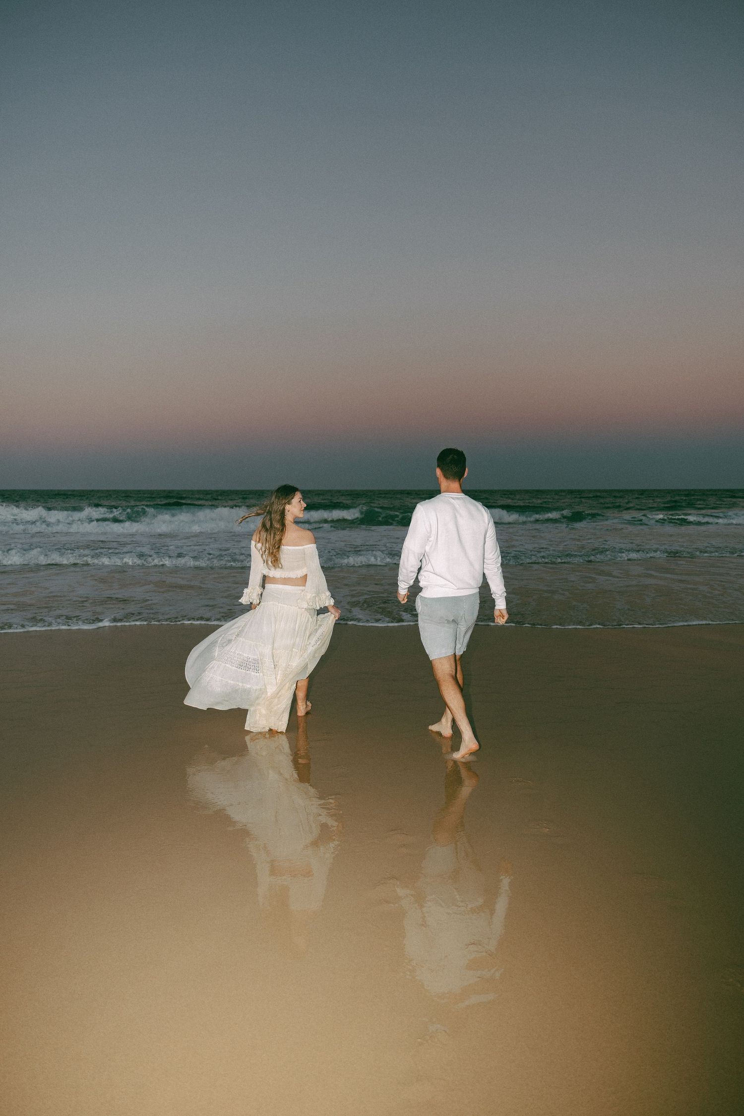 Couple in white clothing walking barefoot toward the ocean on a reflective beach under a pink and blue dusk sky.