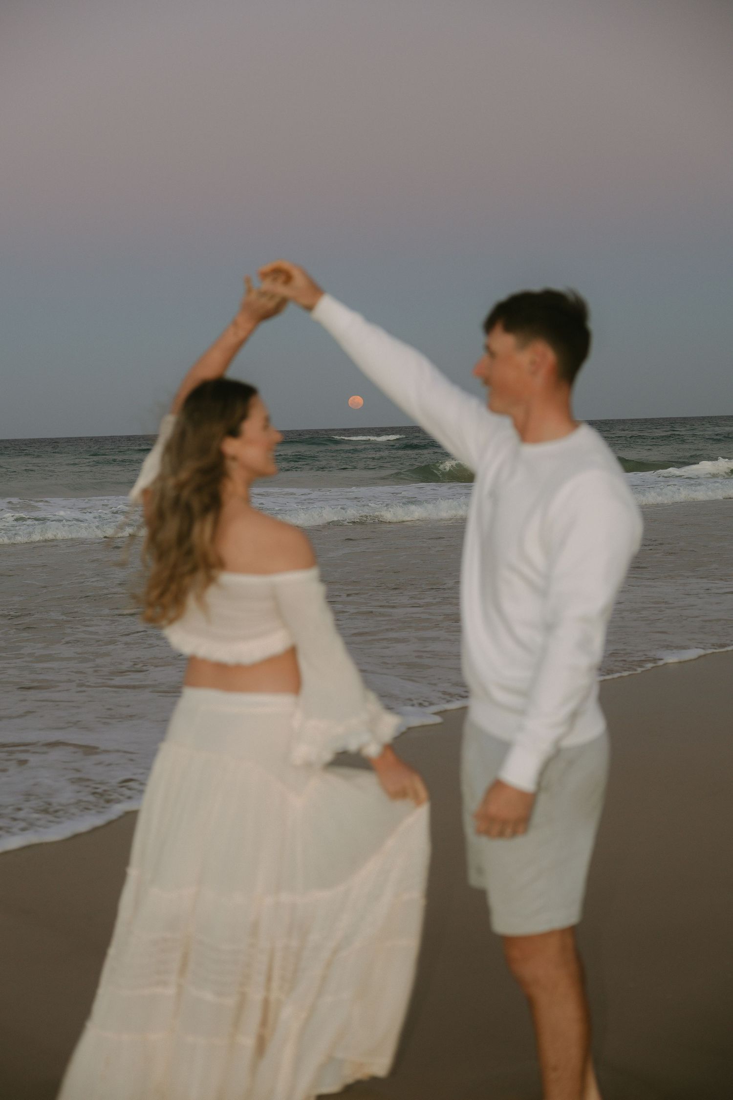 Man twirling a woman in a white boho outfit on the beach at dusk with the moon rising over the ocean.