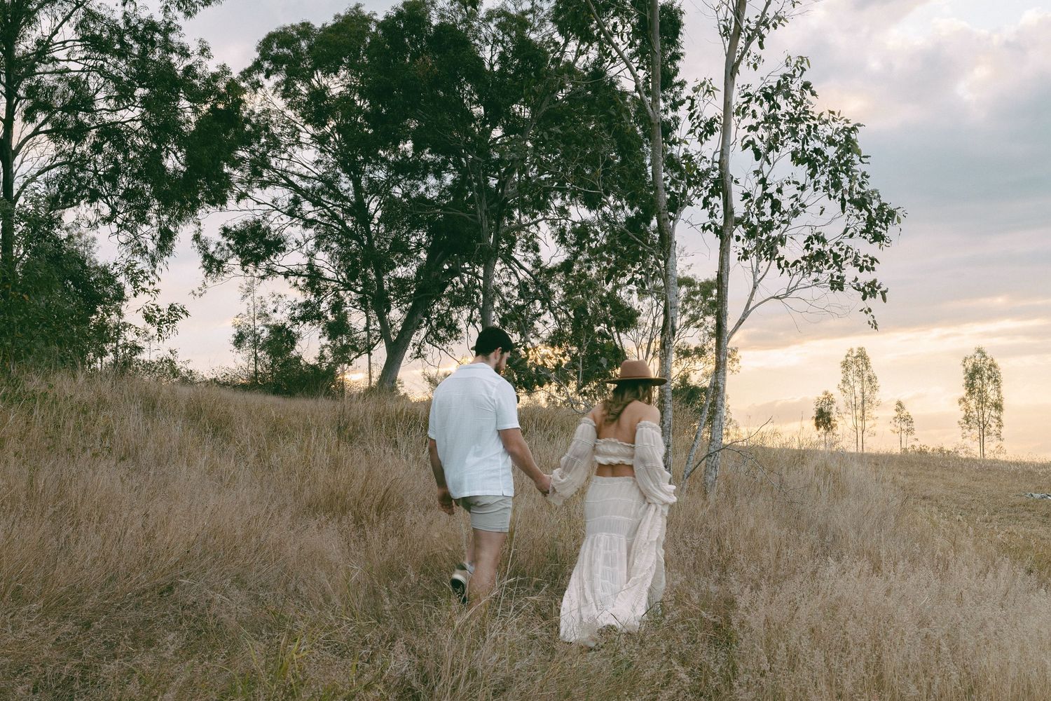 Couple in white outfits holding hands and walking through a golden grass field with gum trees at sunset.