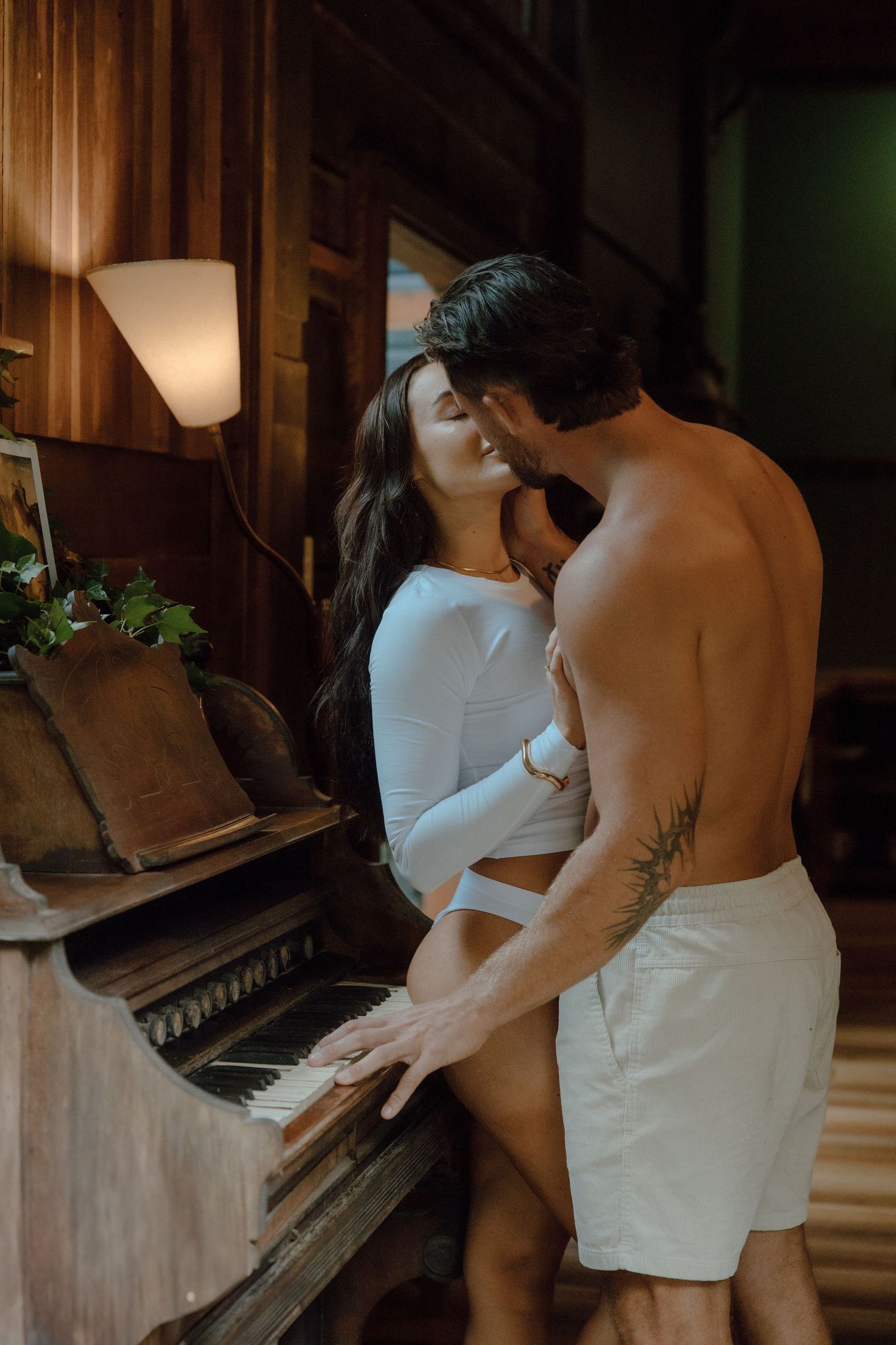 Couple embraces and kisses beside a vintage upright piano in a warmly lit wooden room, the man shirtless and tattooed.