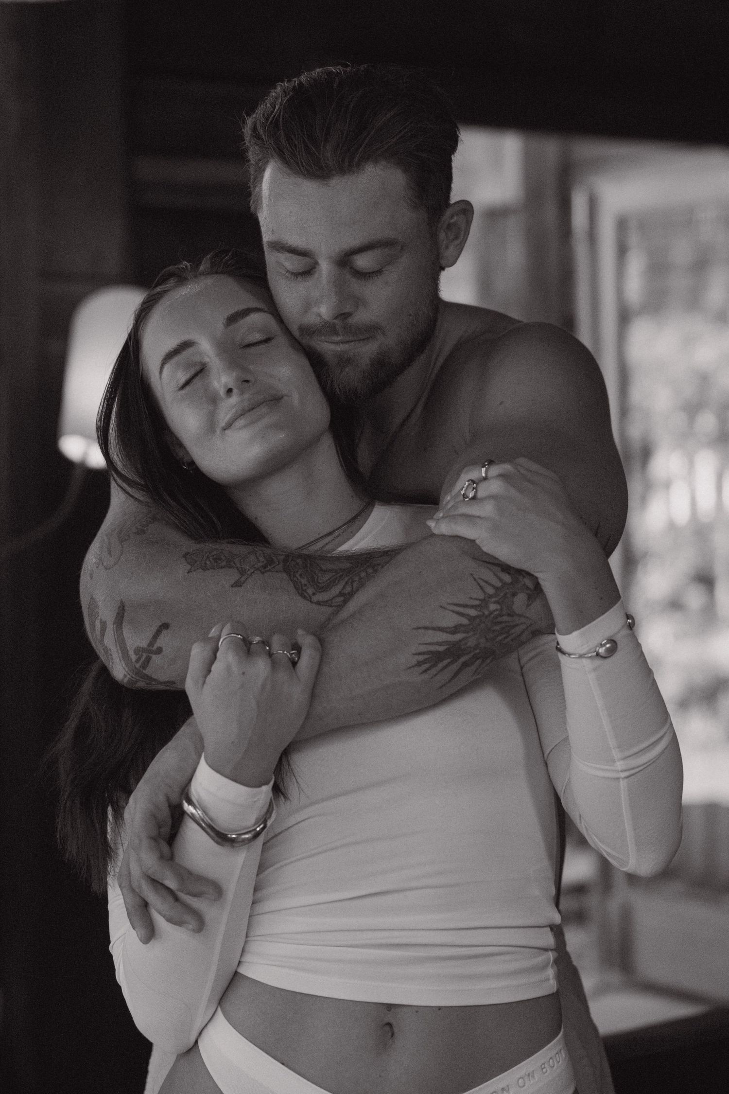 Black and white portrait of a smiling woman with closed eyes held in a tender embrace by a tattooed shirtless man.
