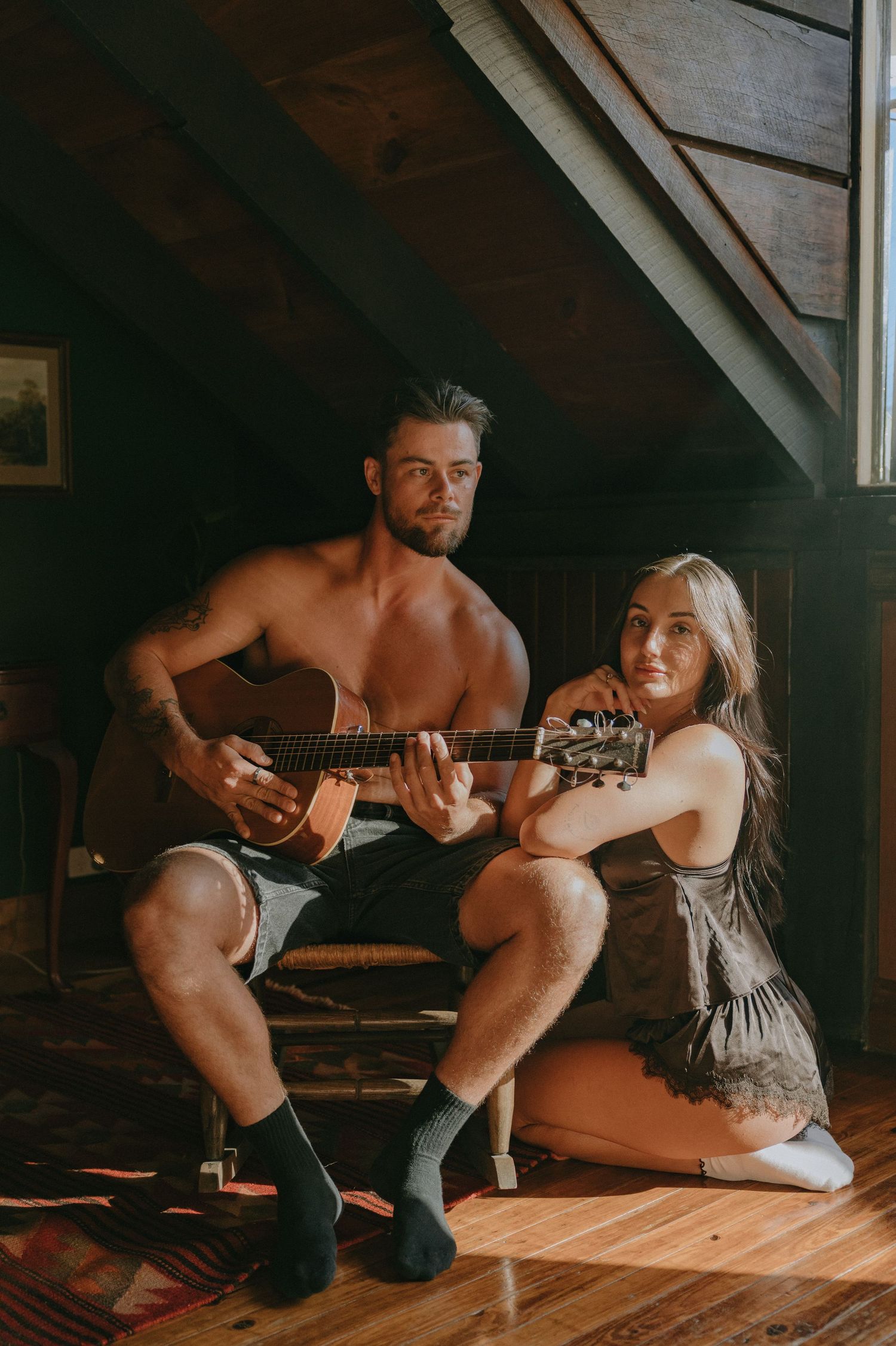 Shirtless man playing acoustic guitar on a wooden chair beside a woman in a lace slip dress in a sunlit rustic attic.