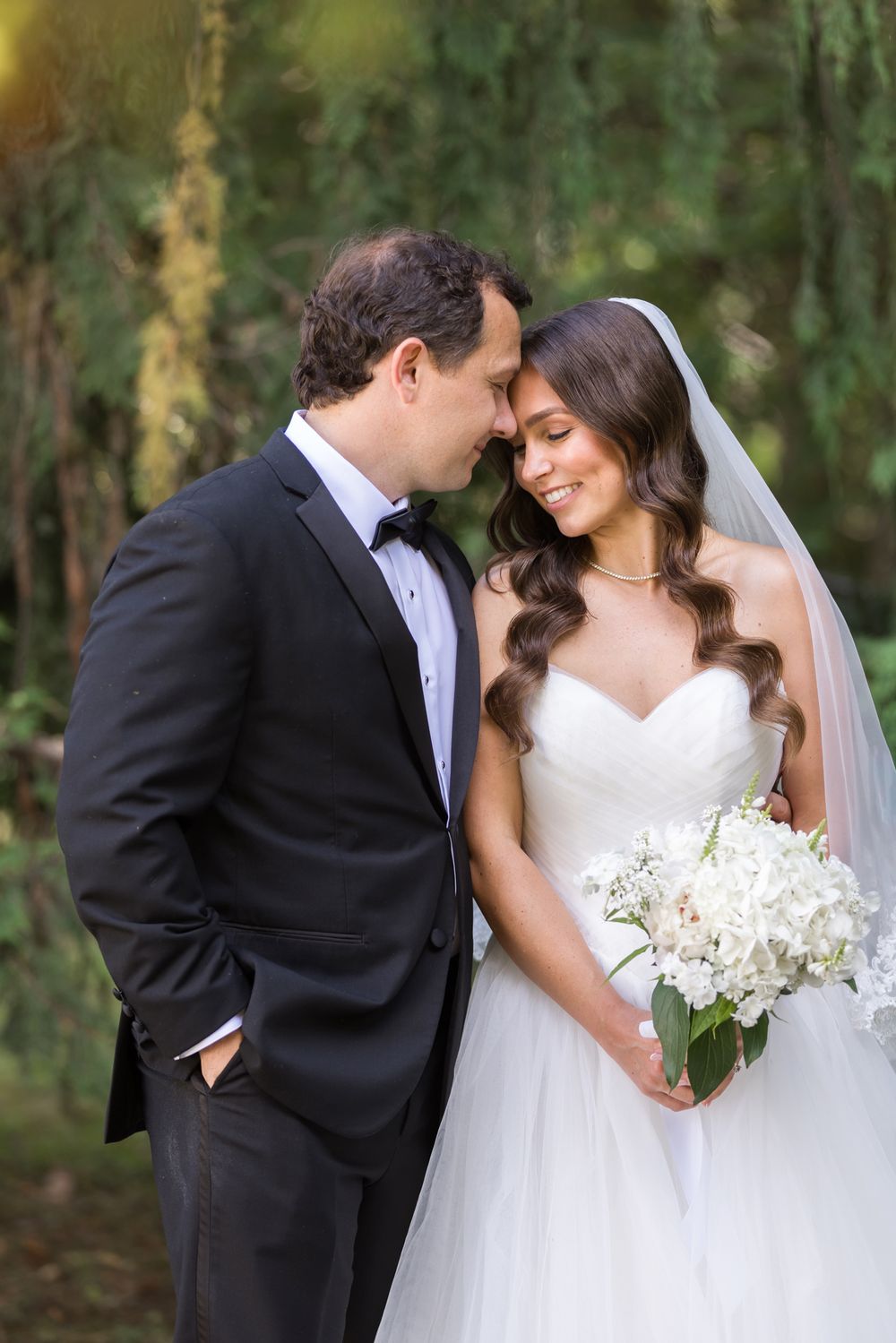 Smiling bride in strapless white gown and veil holds white bouquet as groom in black tuxedo leans in close in front of evergreen.